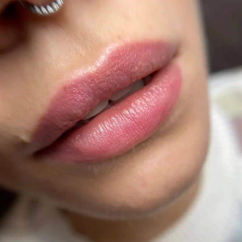 Close-up of a person's lips with pink permanent make up on lips, wearing a hoop earring, on a blurred background.